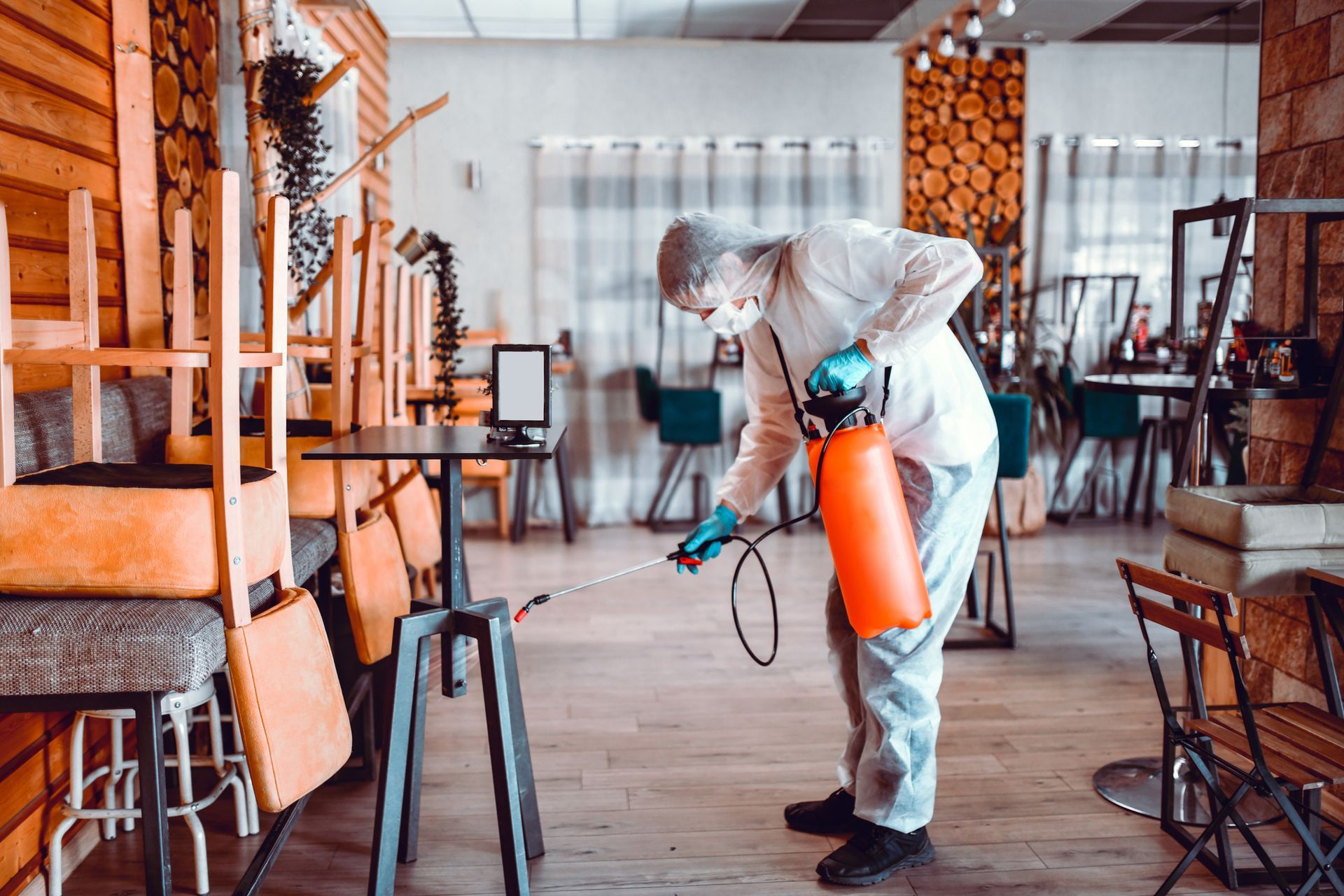 Person spraying disinfectant in a chair-filled room with stacked furniture and curtains