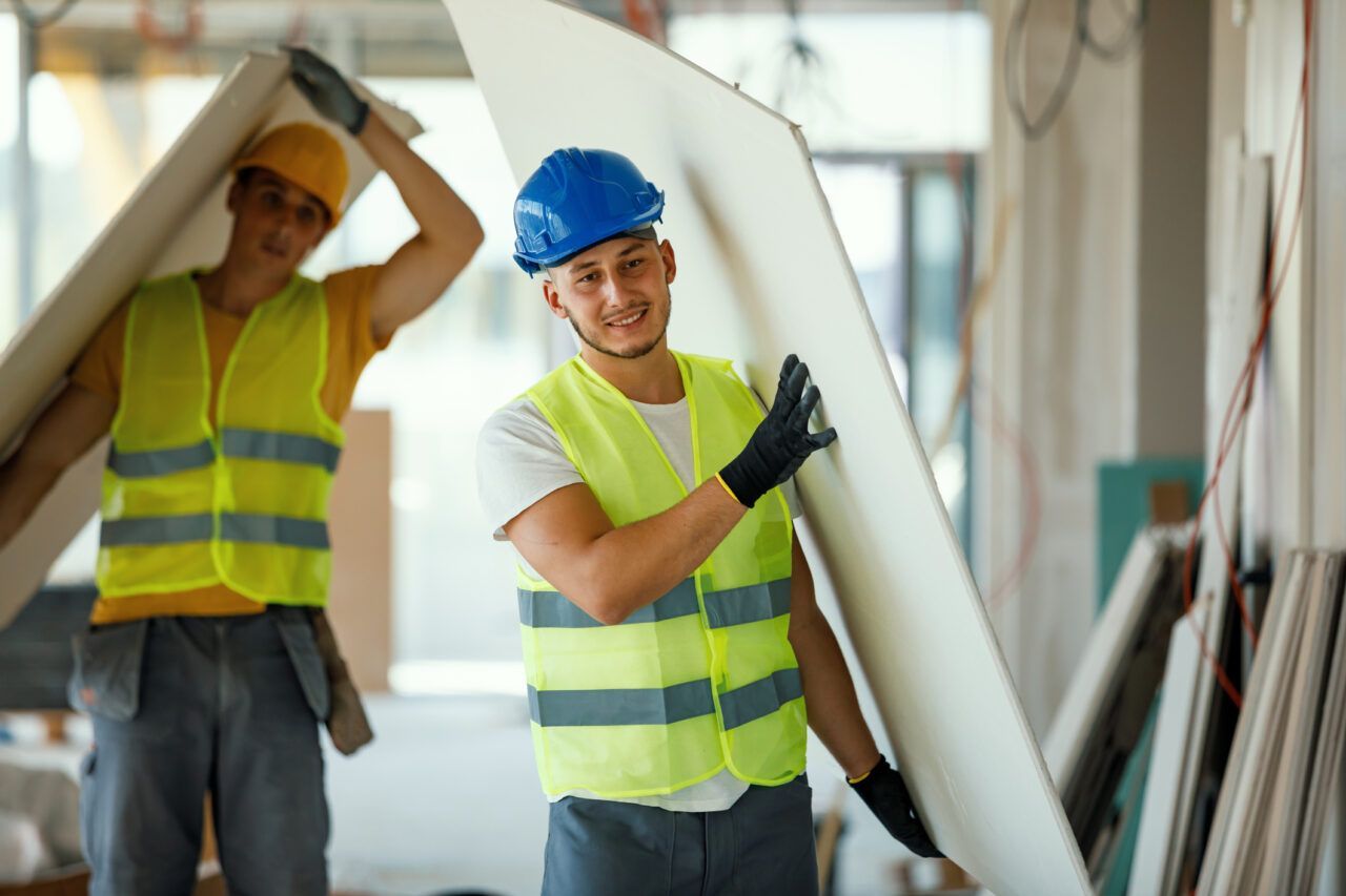 Two construction workers in safety vests carrying a large white pipe inside a building under construction