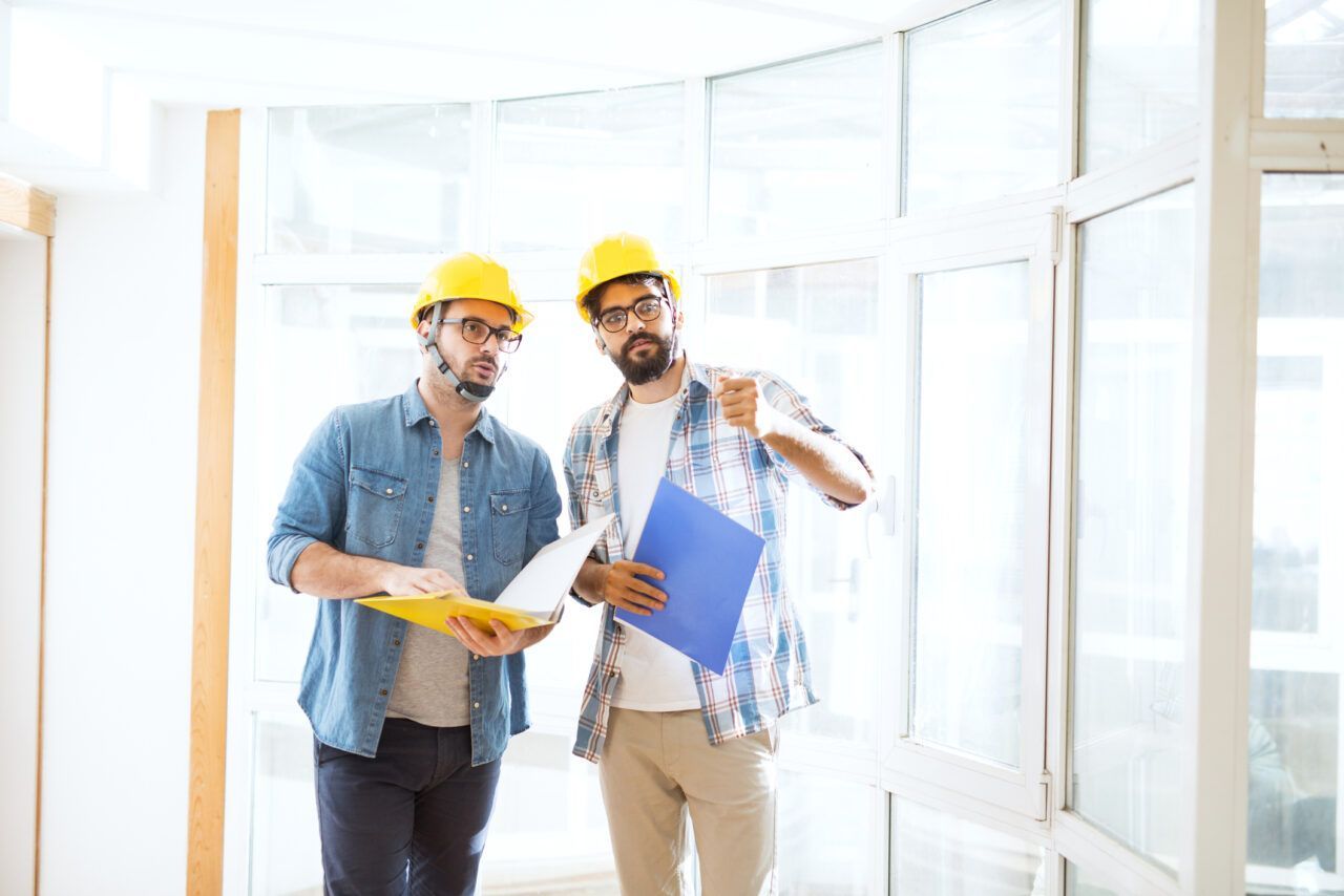 Two construction workers in hard hats reviewing plans inside a bright room