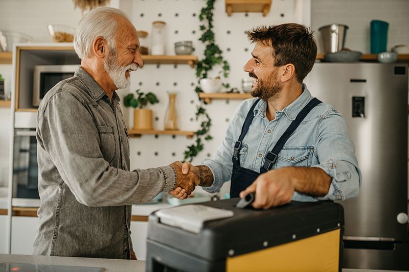 Two men shake hands across a shop counter, smiling in a bright café.