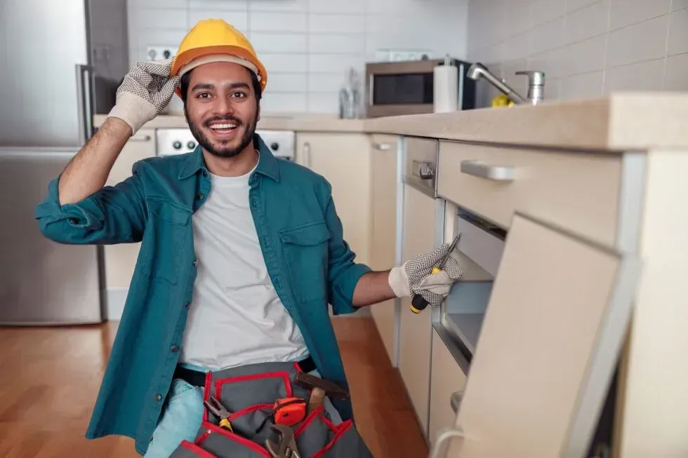 Technician in teal shirt and hard hat smiles while working on kitchen cabinetry with tools.
