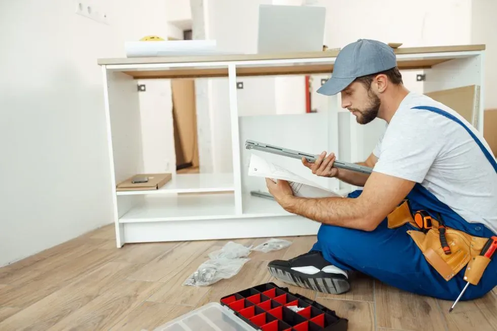 Person assembling white cabinet with a power drill in a room; tool box on the floor.