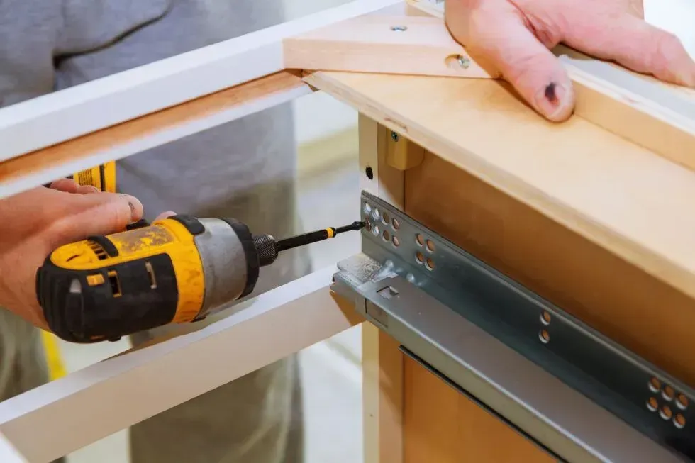 Drill driving a screw into a metal drawer slide on a wooden cabinet frame
