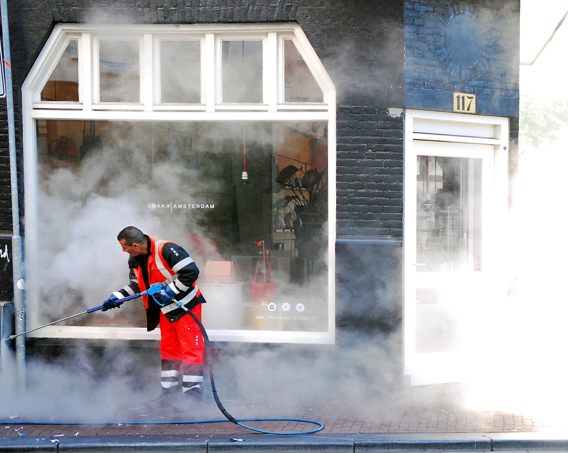 Worker in orange safety gear spraying water on a building facade amid heavy mist and steam.