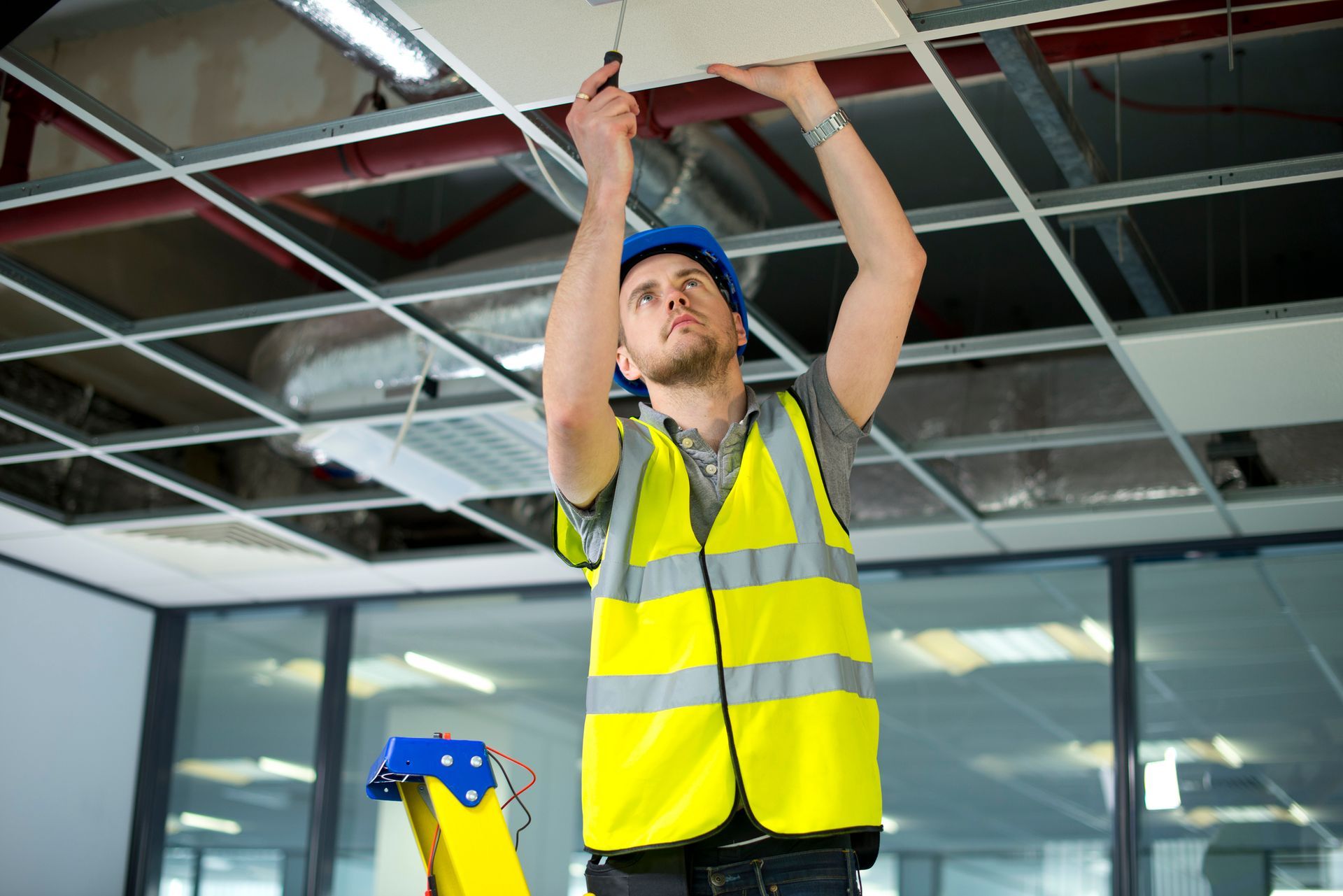 Worker in a hard hat installing ceiling panels in a room with exposed framing and a ladder