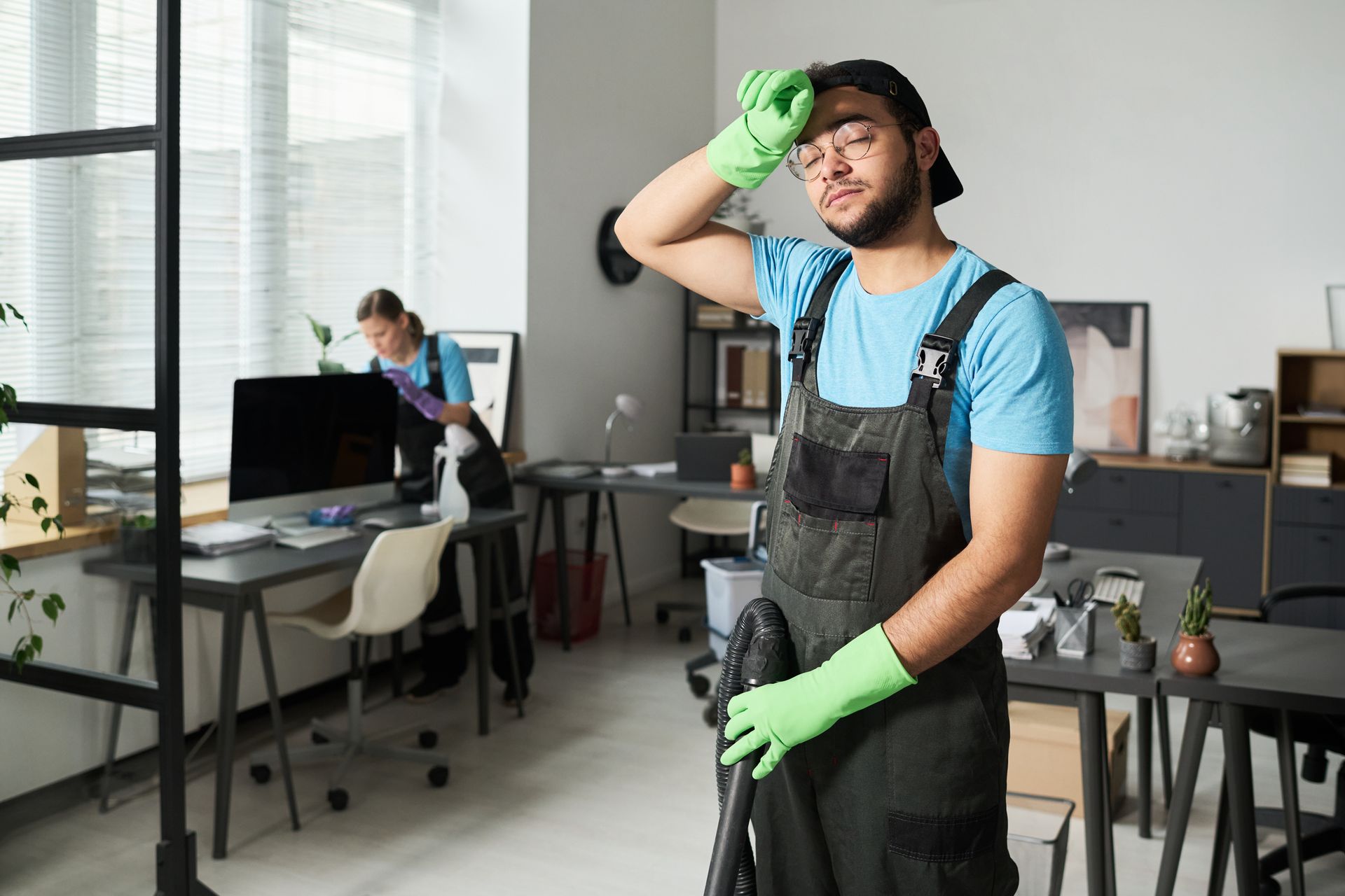 Worker in blue shirt and green gloves pauses in a workshop, holding a tool and wiping his forehead.