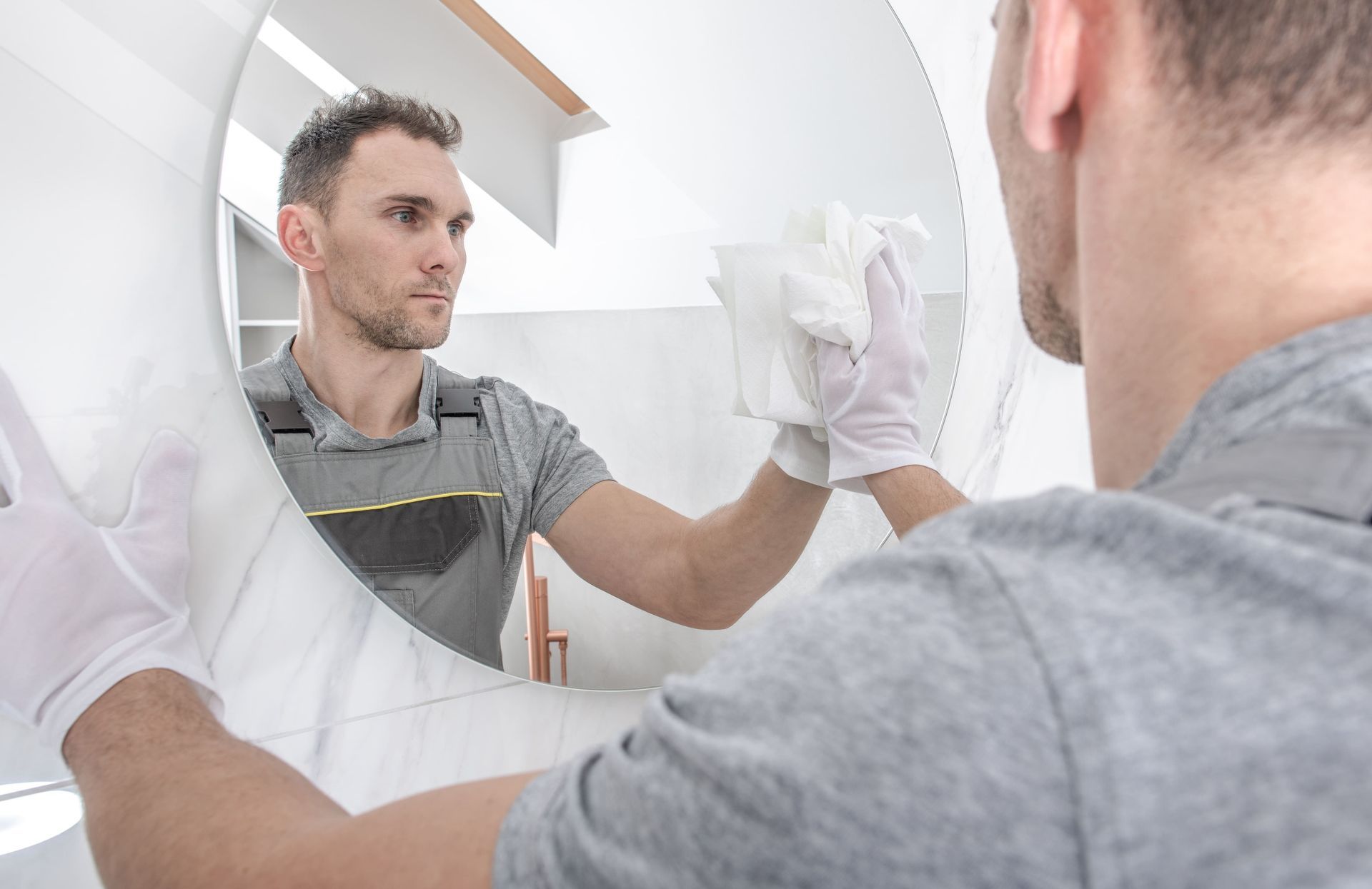 company of house cleaning A man is cleaning a round mirror in a bathroom.