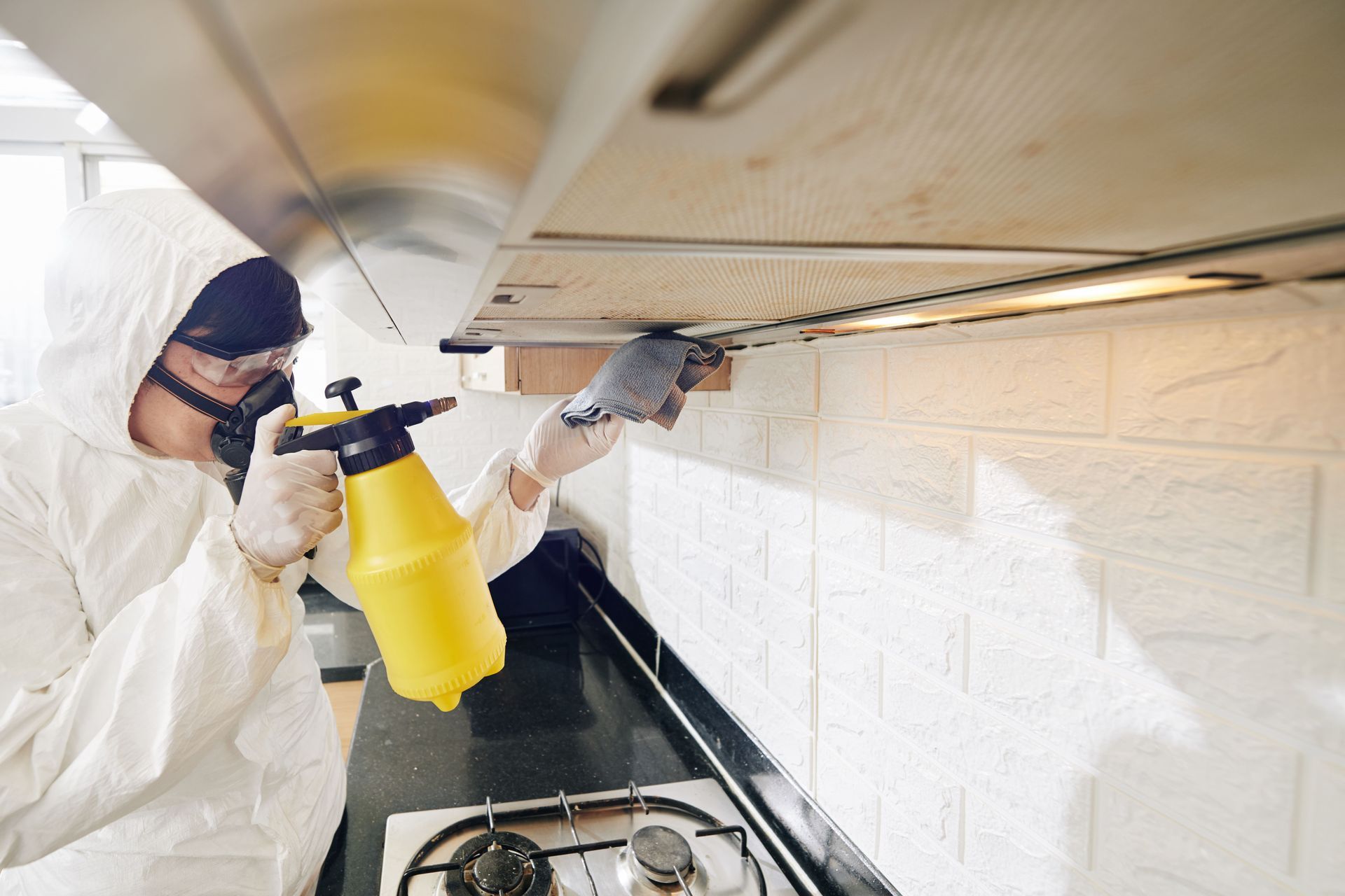 A man in a protective suit is spraying a kitchen hood with a spray bottle.