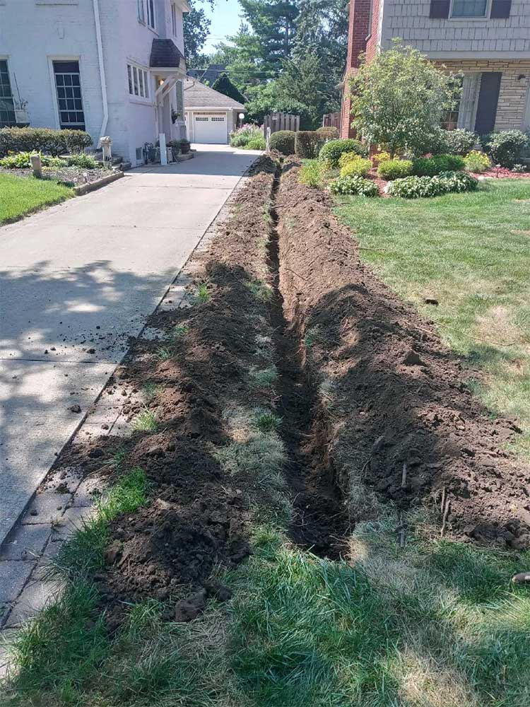 Long trench dug beside a driveway and lawn in a suburban yard, likely for utility work.