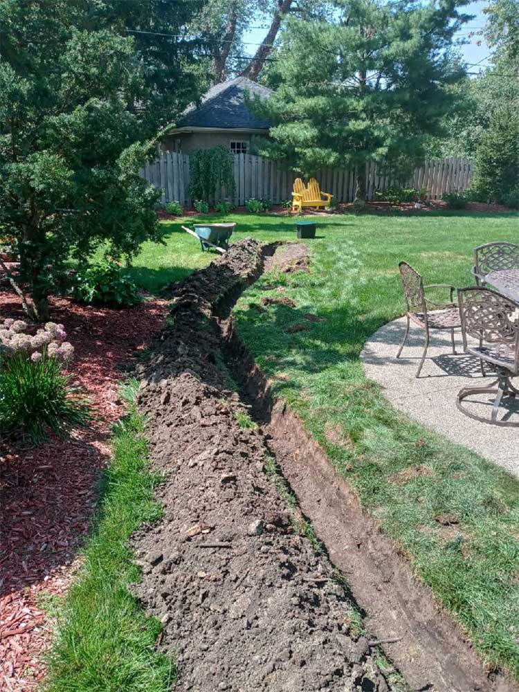 Backyard trench along lawn beside patio chairs and trees, with a shed in the background