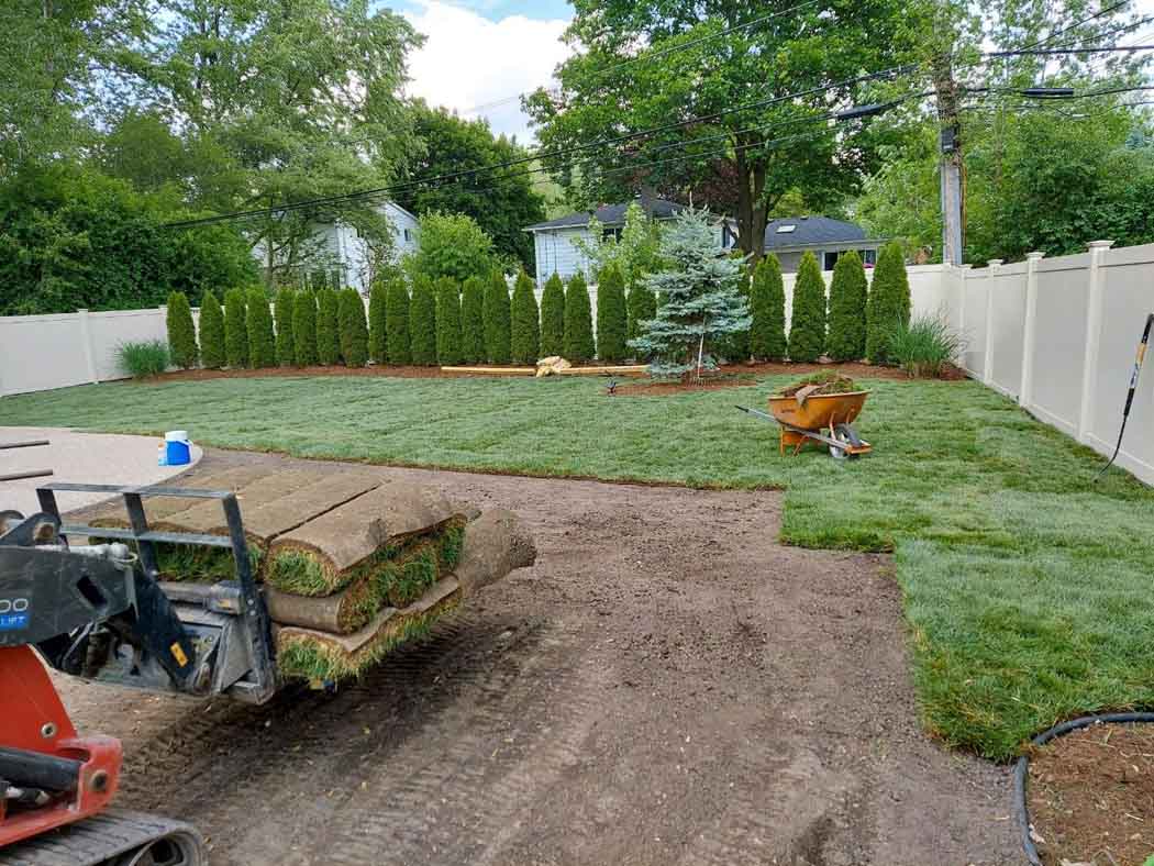 Backyard landscaping with sod, dirt, and a small tractor near a white fence and trees