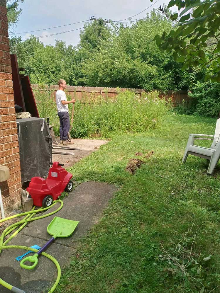 Backyard with overgrown grass, a person standing near the fence, and children’s toys on the patio.