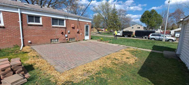 Backyard patio with gray pavers beside a red brick house, bordered by grass and a small stack of bricks