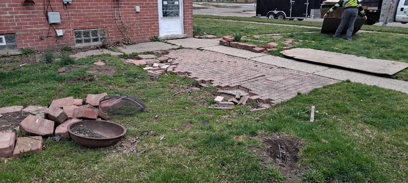 Broken walkway beside a brick house with scattered stones and muddy grass