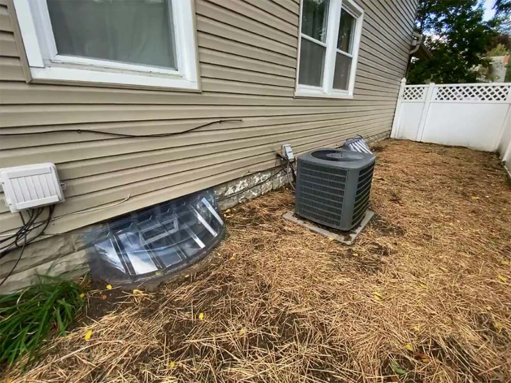 Side of beige house with air conditioner unit and covered basement window in a mulched yard, beside white fence