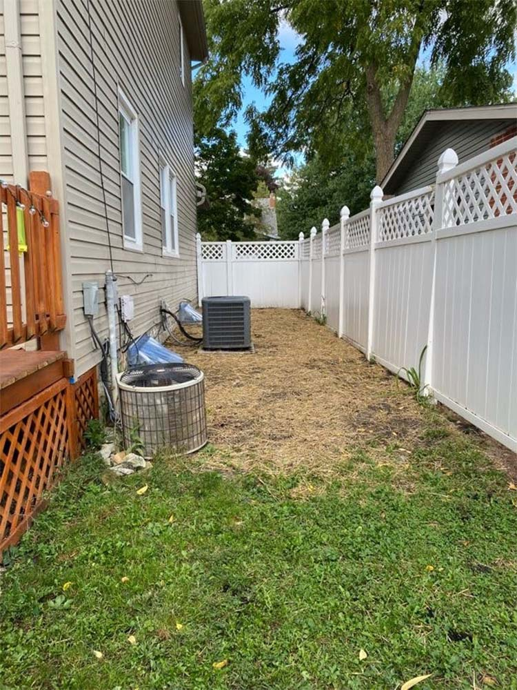 Narrow side yard with white fence, beige house siding, grass, and a small metal container near the wall