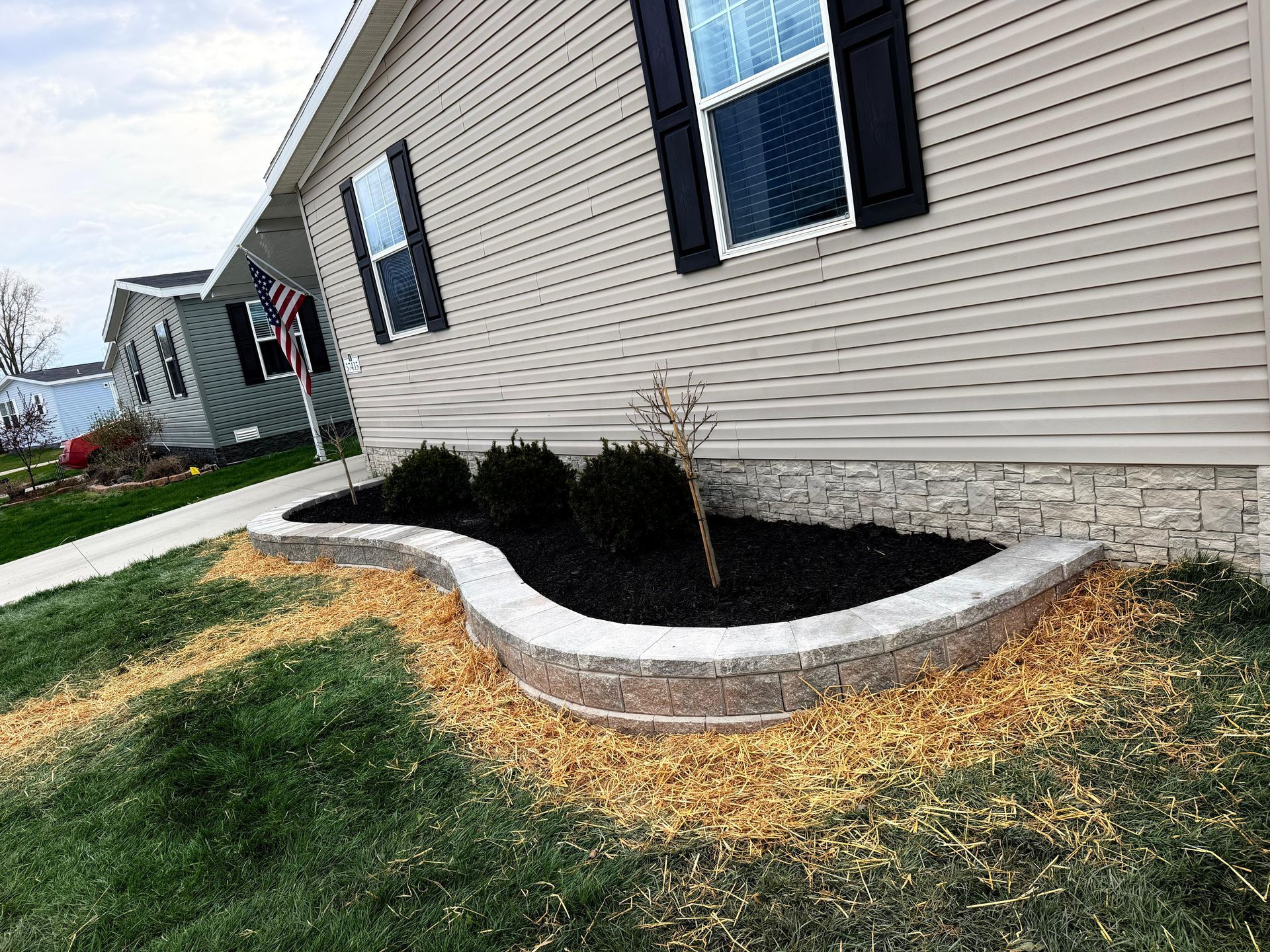 Curved stone flower bed with black mulch and small shrubs beside a beige house with blue shutters