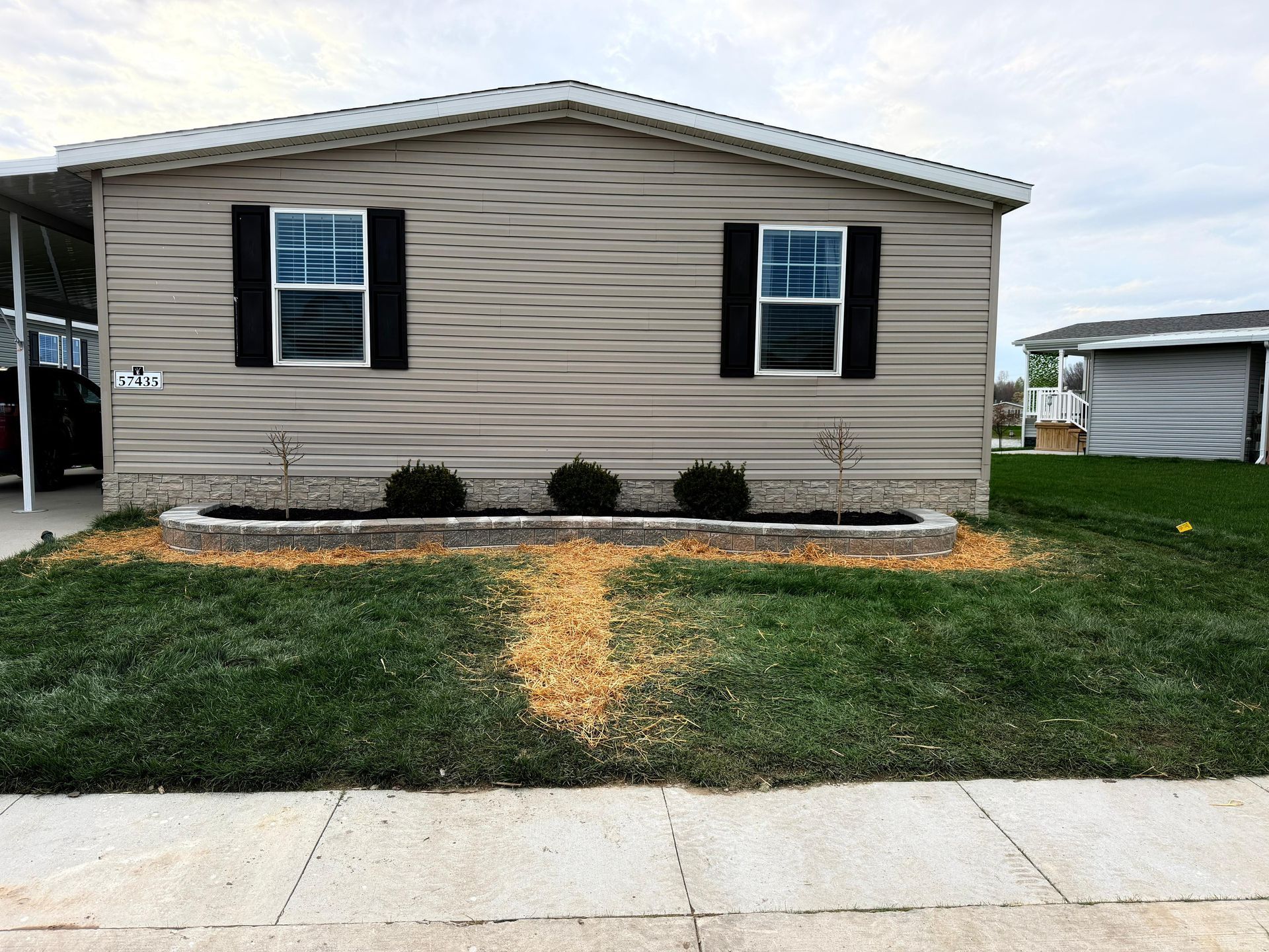 Single-story beige mobile home with black shutters, front lawn, and a dirt path leading to the entrance
