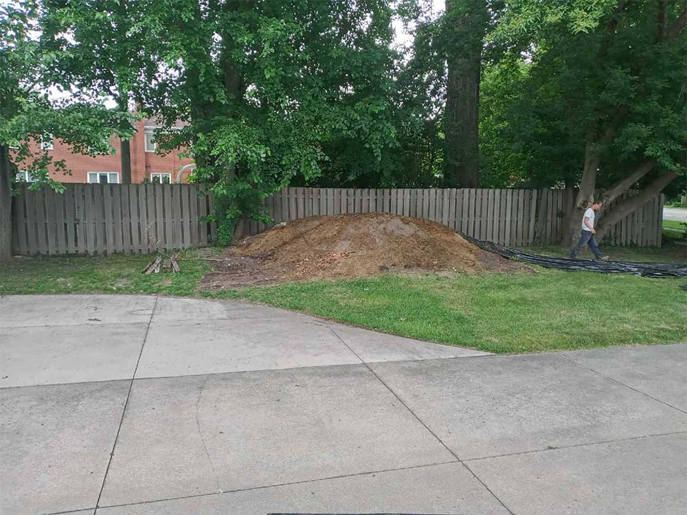 Backyard with a dirt mound by a wooden fence, grassy lawn, and a concrete driveway in the foreground