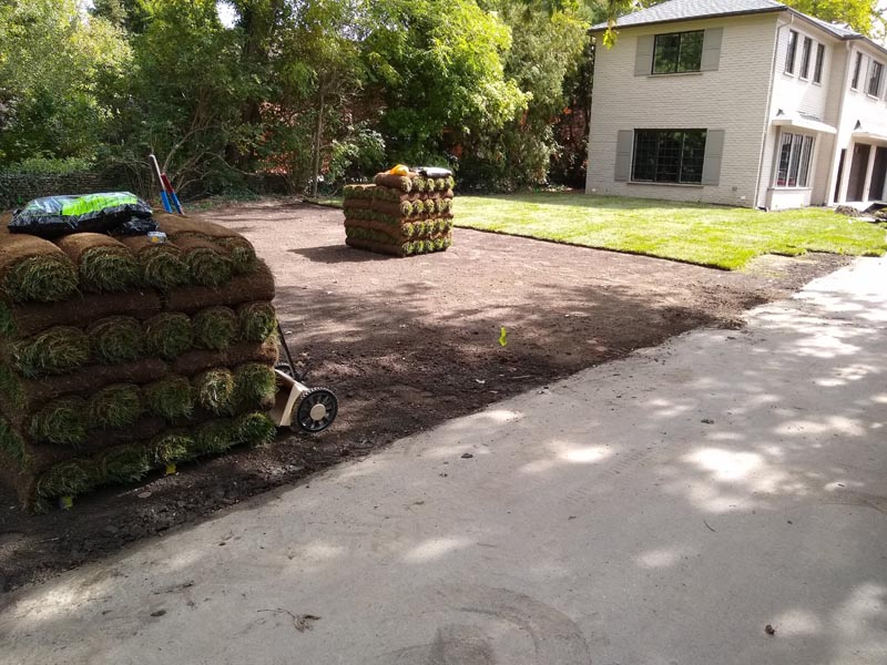Landscaped yard with fresh mulch, stacked sod, and a white house beside a driveway.