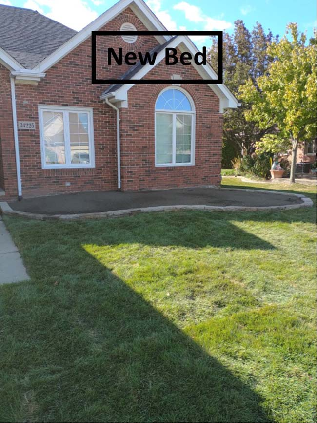 Brick house front yard with a black labeled sign reading “New Bed” over a garden area