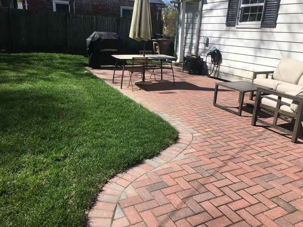 Backyard patio with red brick walkway, green lawn, grill, table, and chairs beside a house