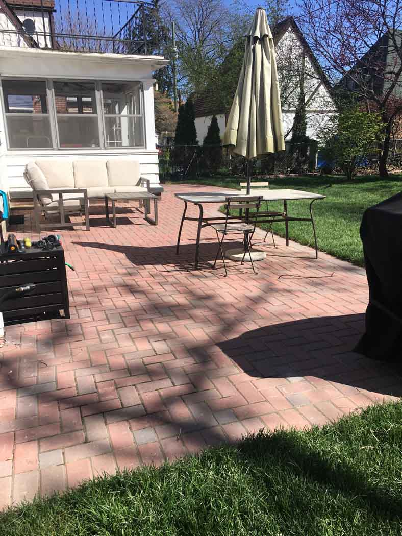 Backyard patio with brick pavers, tables and chairs, an umbrella, and a small shed in sunlight.