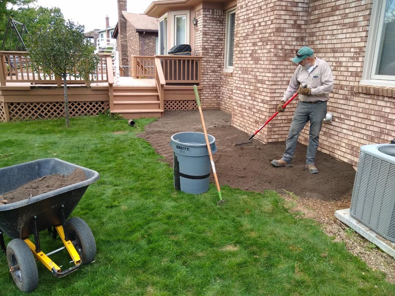Person raking dirt beside a brick house with a wheelbarrow and yard tools on a lawn