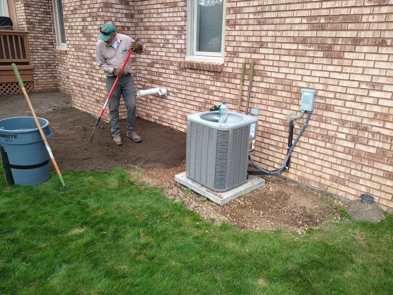 Worker shoveling dirt beside a brick house and outdoor air conditioner unit