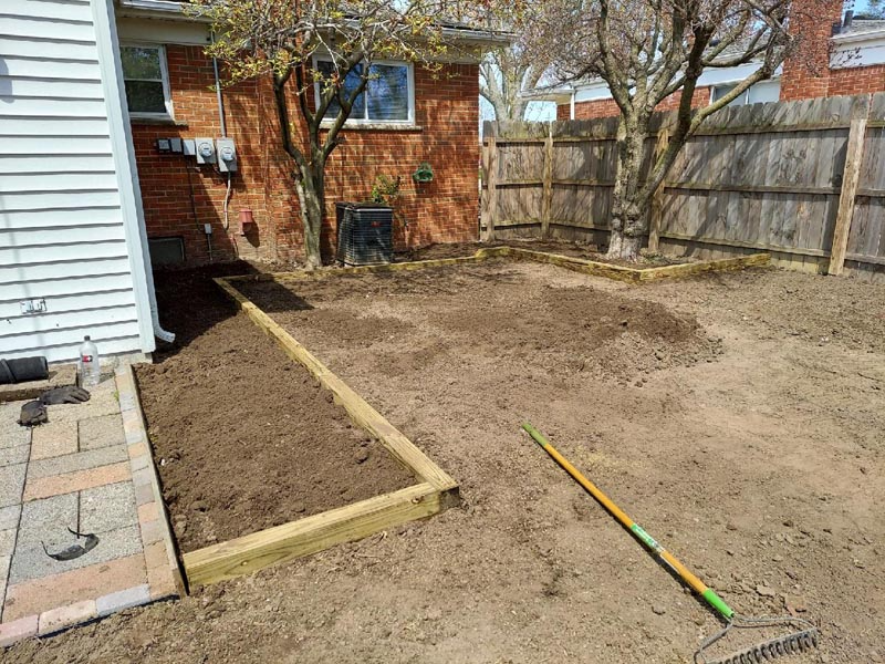 Backyard with fresh soil, wooden border, fence, brick shed, and rake on the ground.