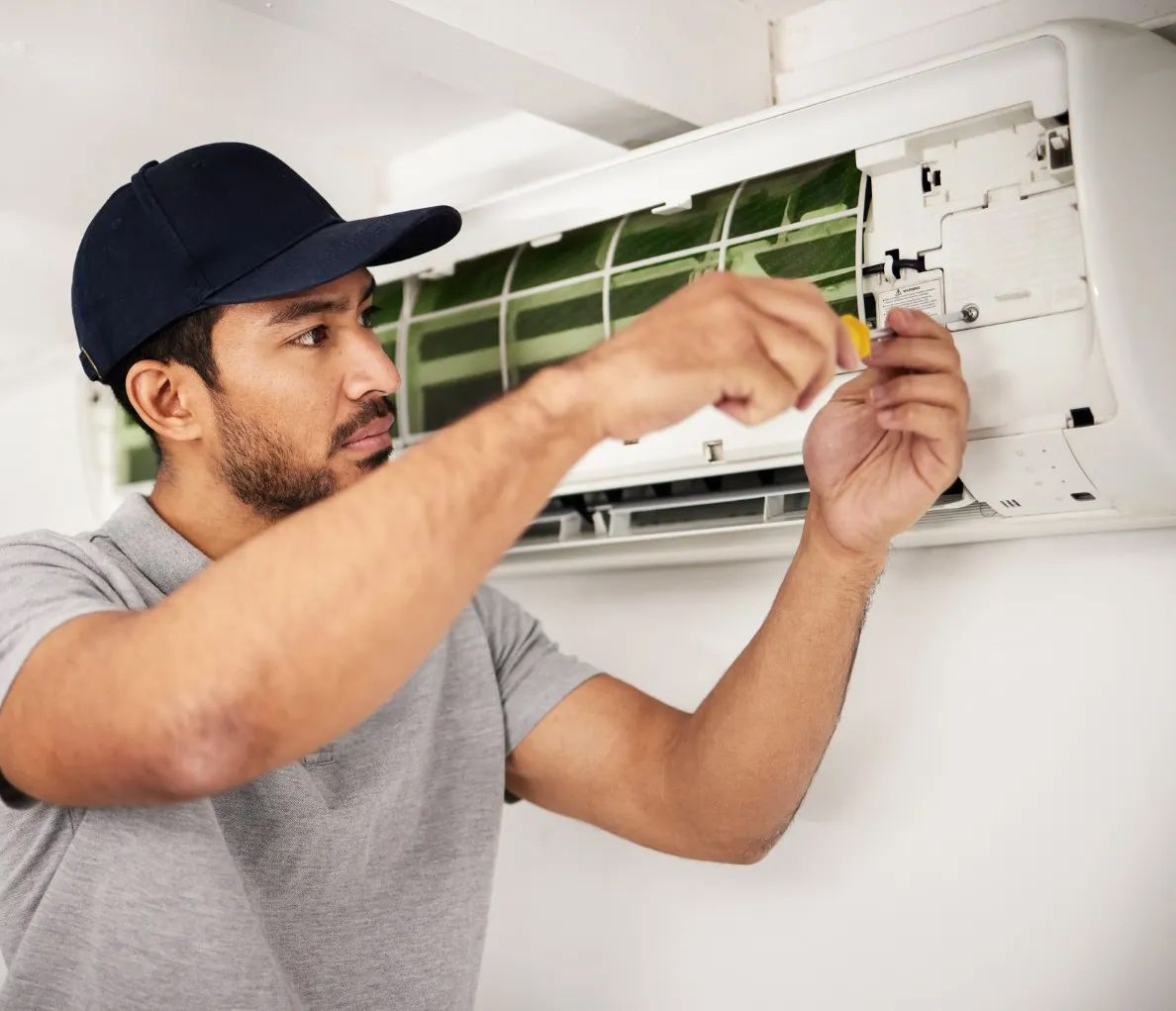 Technician replacing an air conditioner filter indoors, wearing a cap and gray shirt.