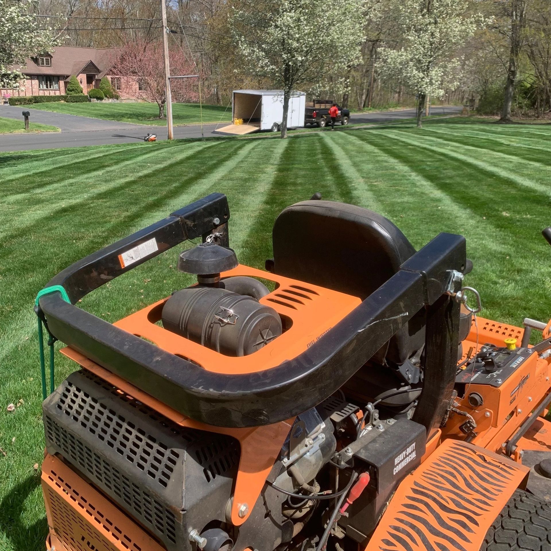 Orange riding mower on striped green lawn with trees and a house in the background