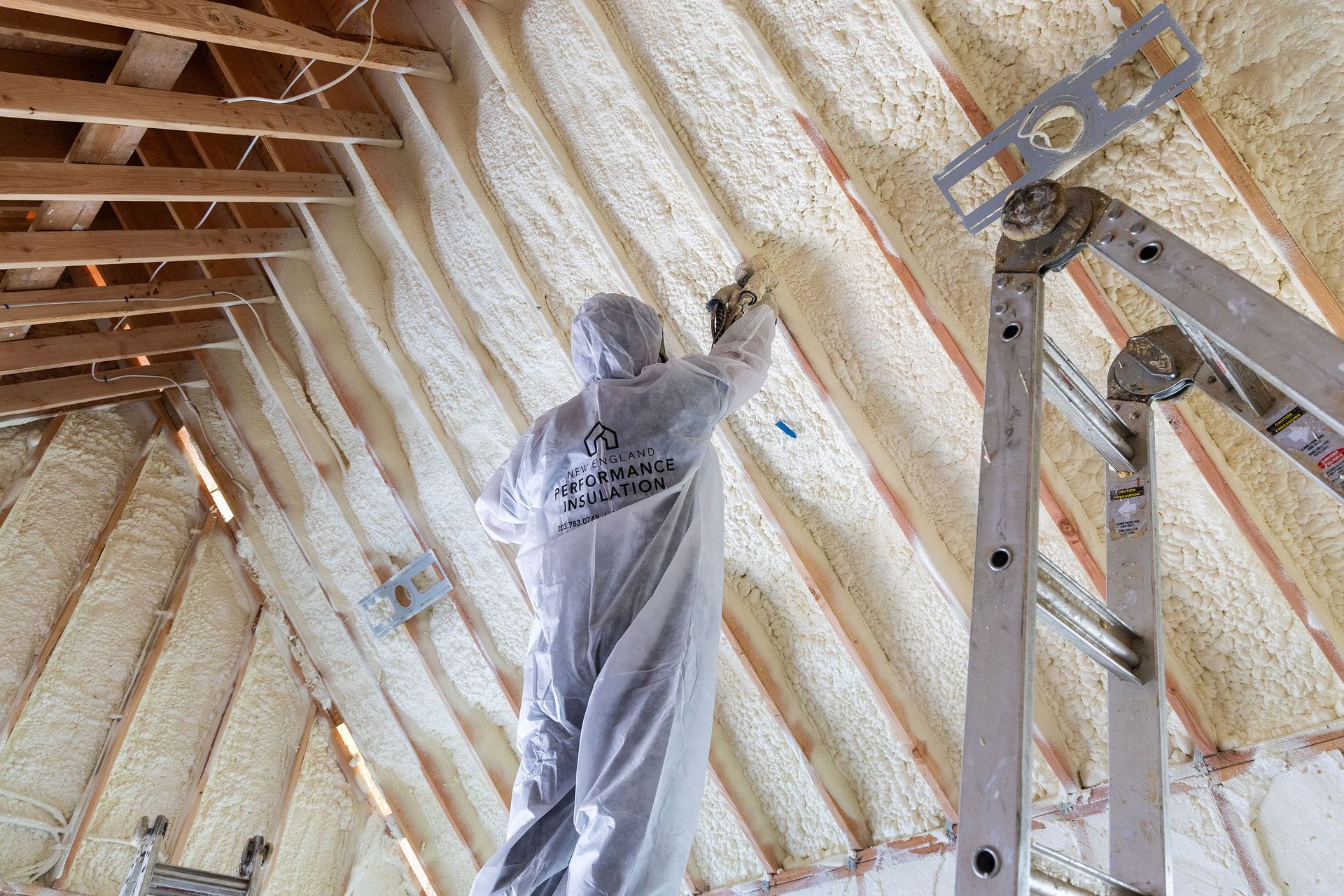 Worker spray-foaming insulation on an attic ceiling beside a ladder