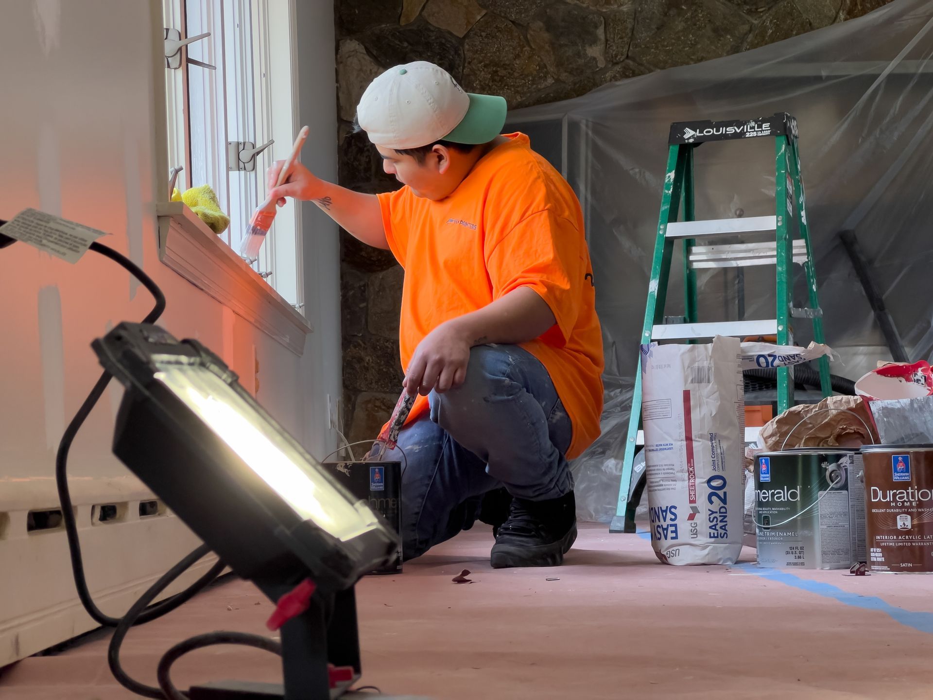 Worker in orange shirt painting a wall inside a room under renovation, with tools and ladder nearby.
