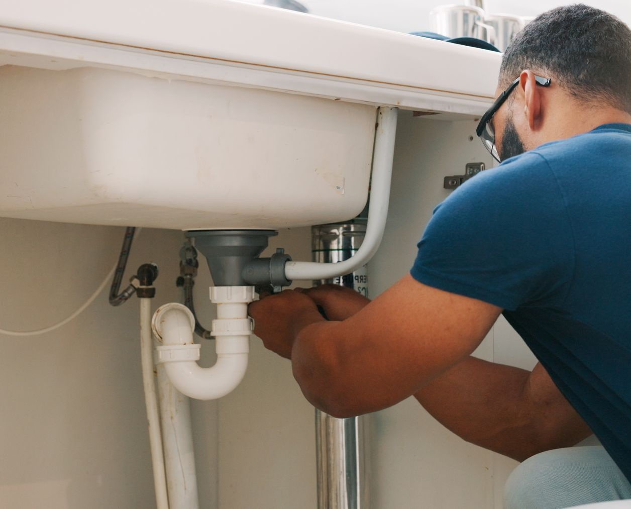 Plumber repairing under-sink pipes beneath a white bathroom sink with tools in hand