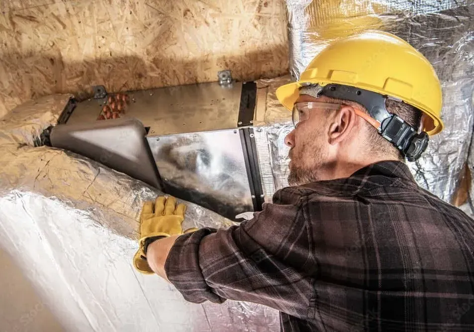 A worker in a yellow hard hat and gloves is installing ductwork in an attic, surrounded by insulation.