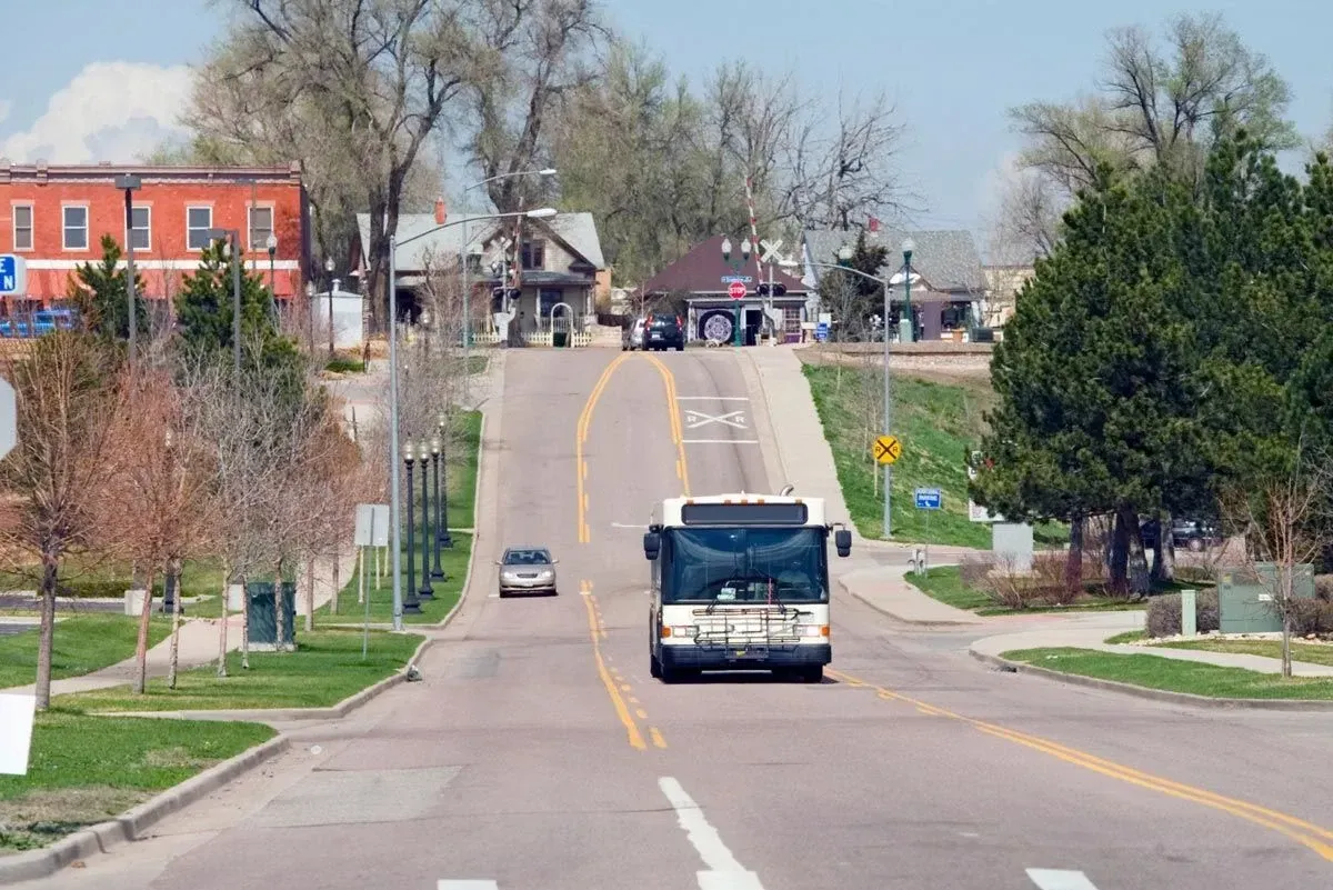 Bus traveling uphill on a two-lane road in a small town; buildings and trees line the street.