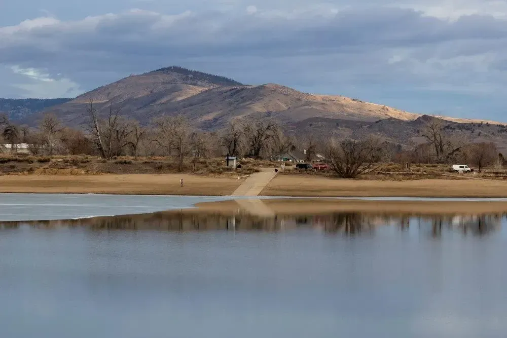 A lake reflects the sky and a distant mountain. A boat ramp leads to a sandy shore with buildings and bare trees.