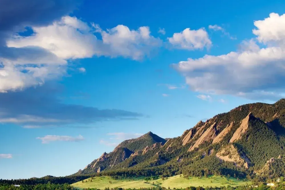 Mountains under a blue sky with fluffy white clouds; green trees on the mountainsides and in the foreground.