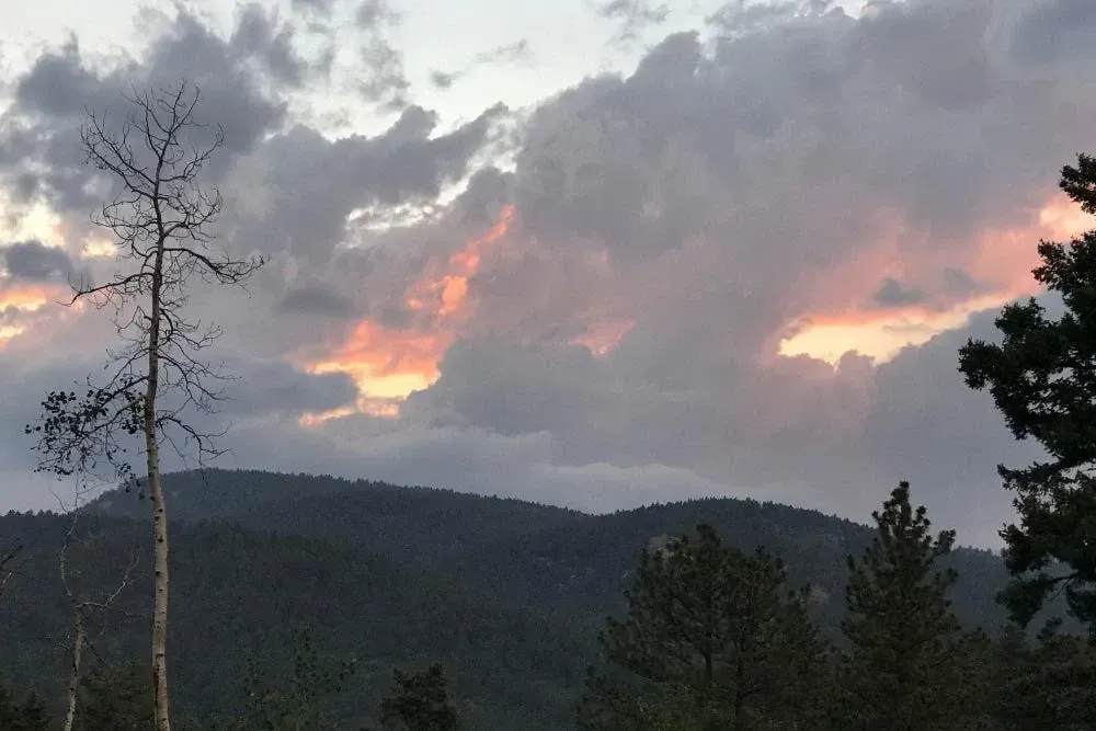 A colorful sunset glows behind dramatic clouds above a dark, silhouetted mountain range and pine trees.