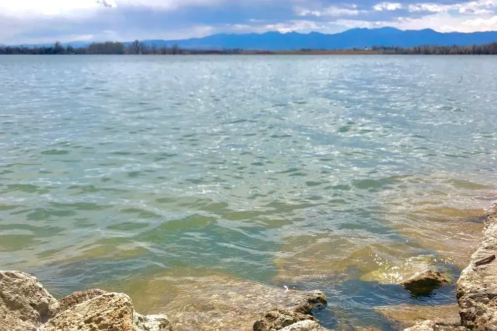 Lake with rippling water under a cloudy sky, mountains in the distance; shoreline with rocks in the foreground.