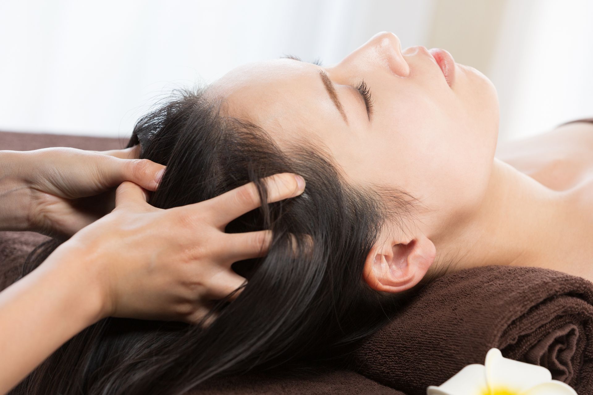 Frau erhält eine Kopfhautmassage, Augen geschlossen, mit den Händen massiert sie ihr Haar. Braunes Handtuch und weiße Blume.