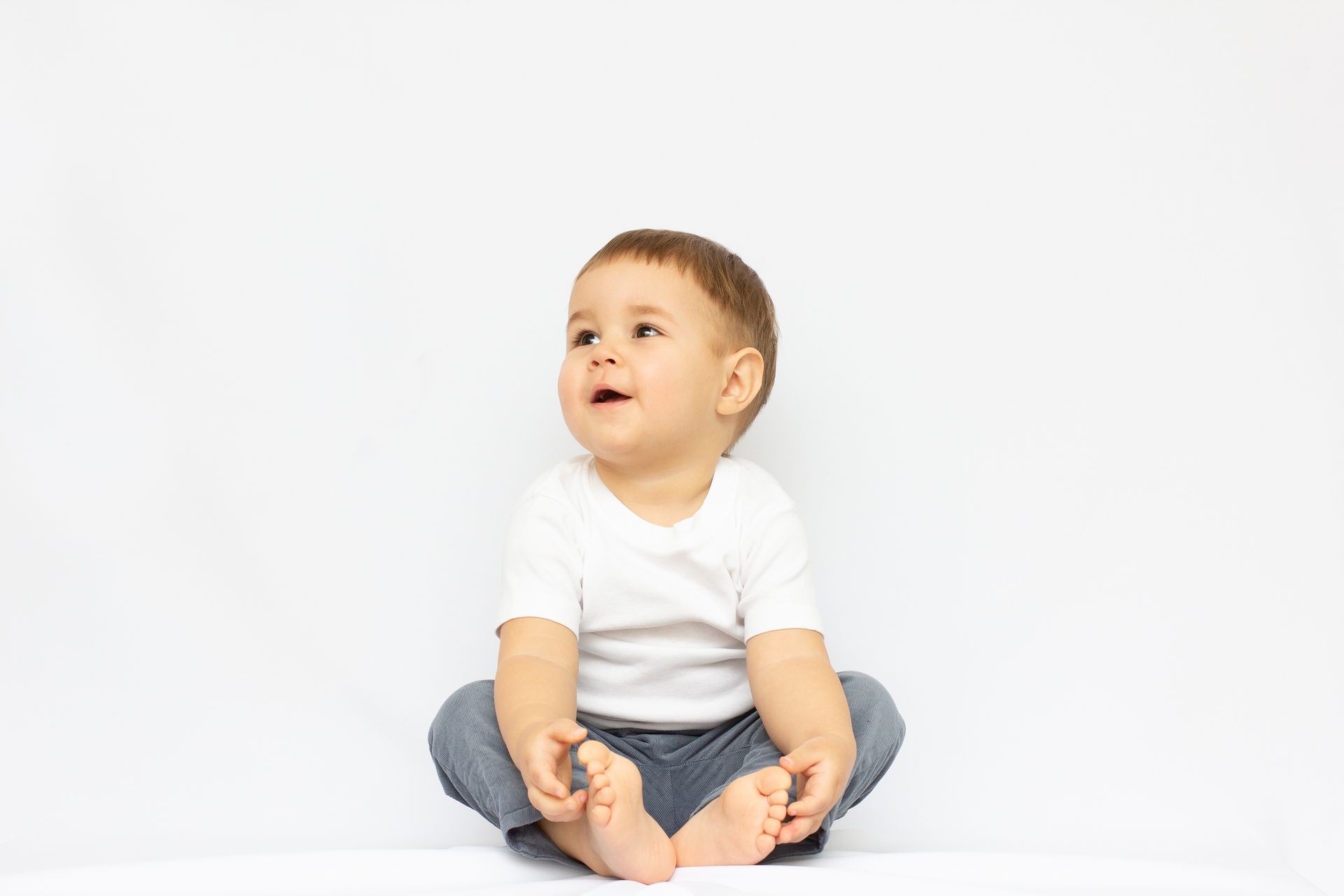 Ein Baby, das ein weißes Hemd und eine graue Hose trägt, sitzt auf einer weißen Fläche und schaut nach oben.