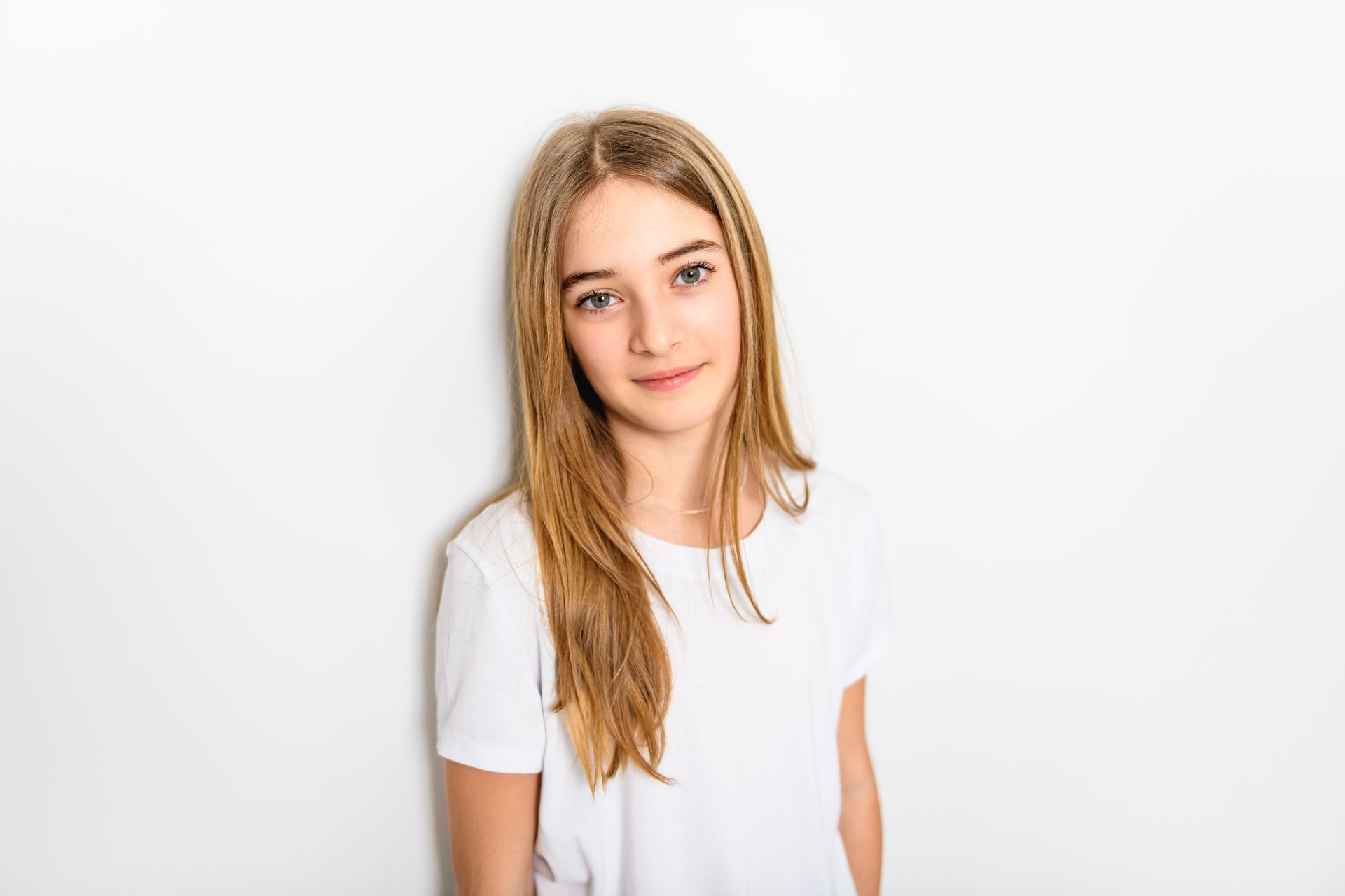 Junges Mädchen mit langen blonden Haaren lächelt vor einer weißen Wand und trägt ein weißes T-Shirt.