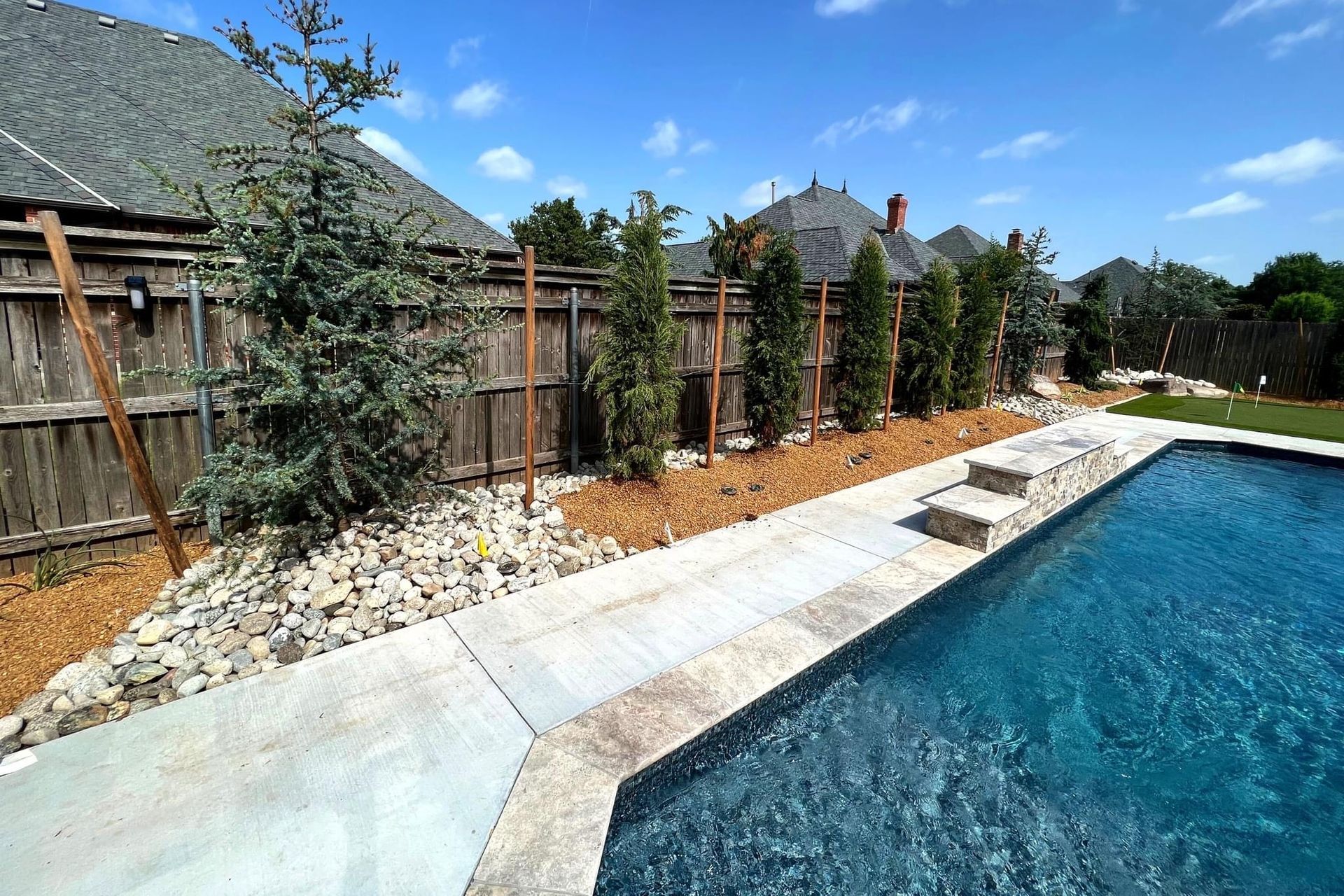 Backyard pool with concrete deck, gravel border, wooden fence, and tall shrubs under a bright sky
