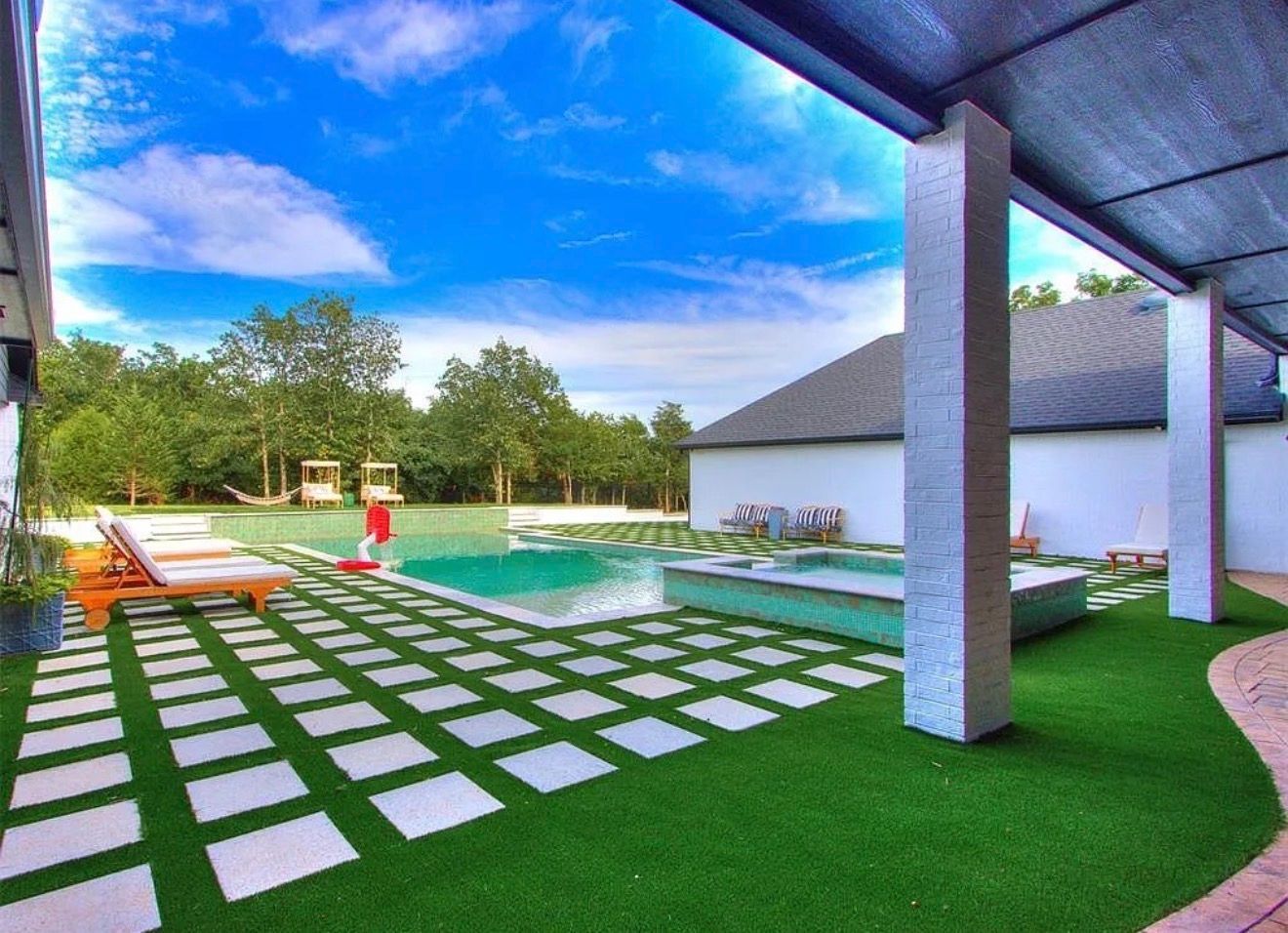Modern backyard pool with stone path, lounge chairs, and covered patio under a blue sky