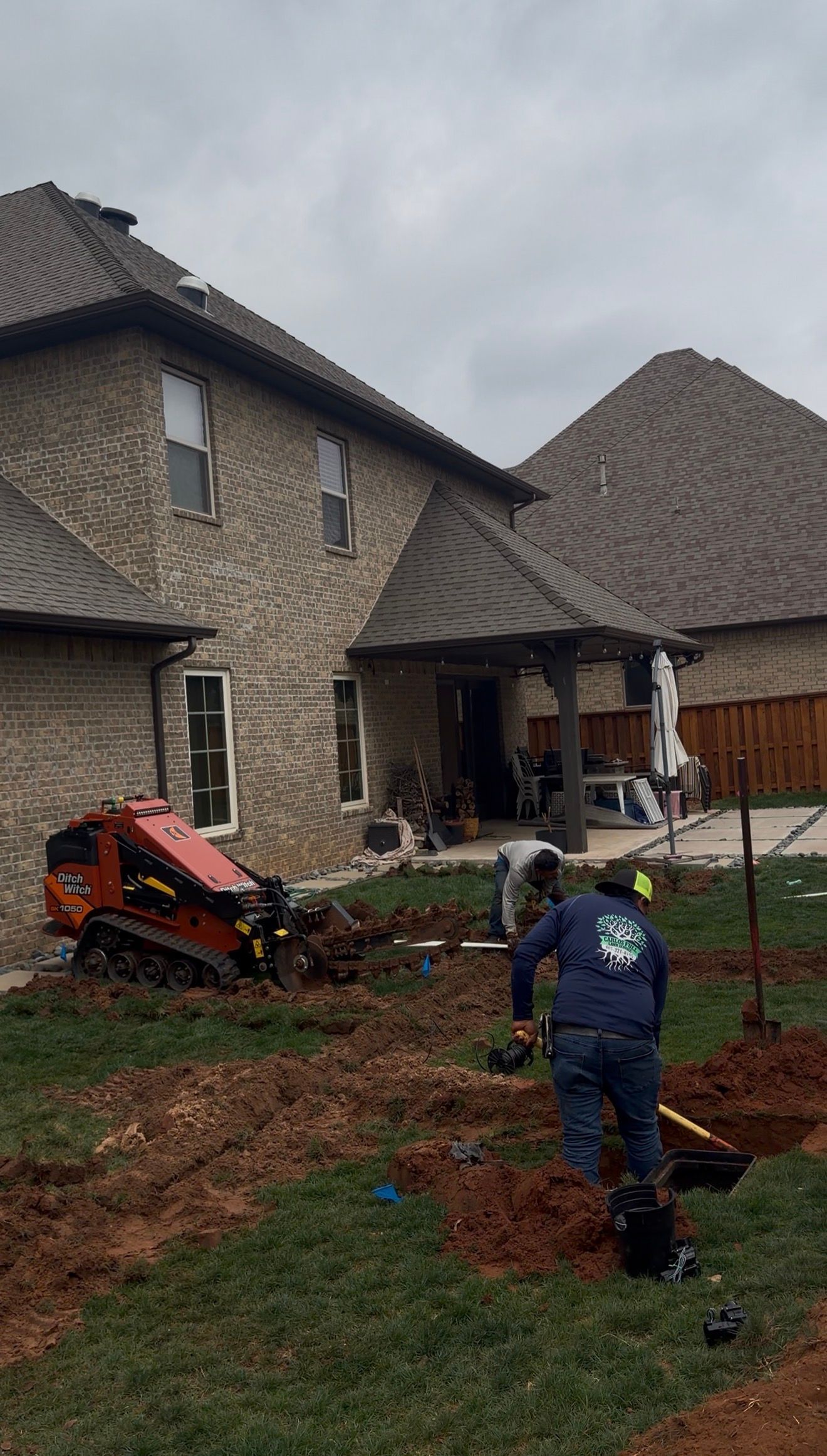 Two workers in a yard installing a small tree beside a stone house and driveway