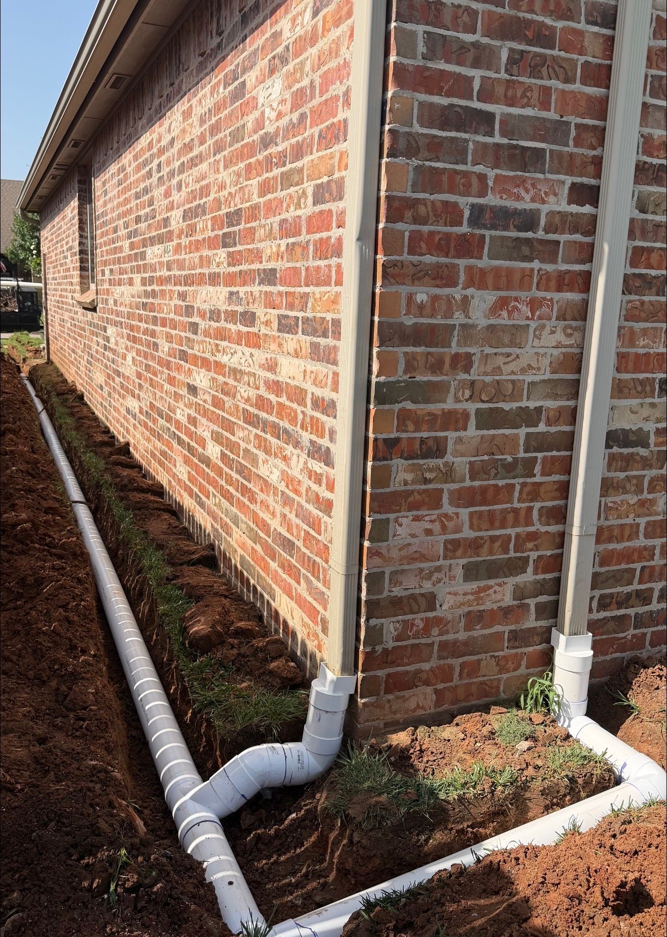Brick house exterior with white drain pipes running along the foundation and down the corner.