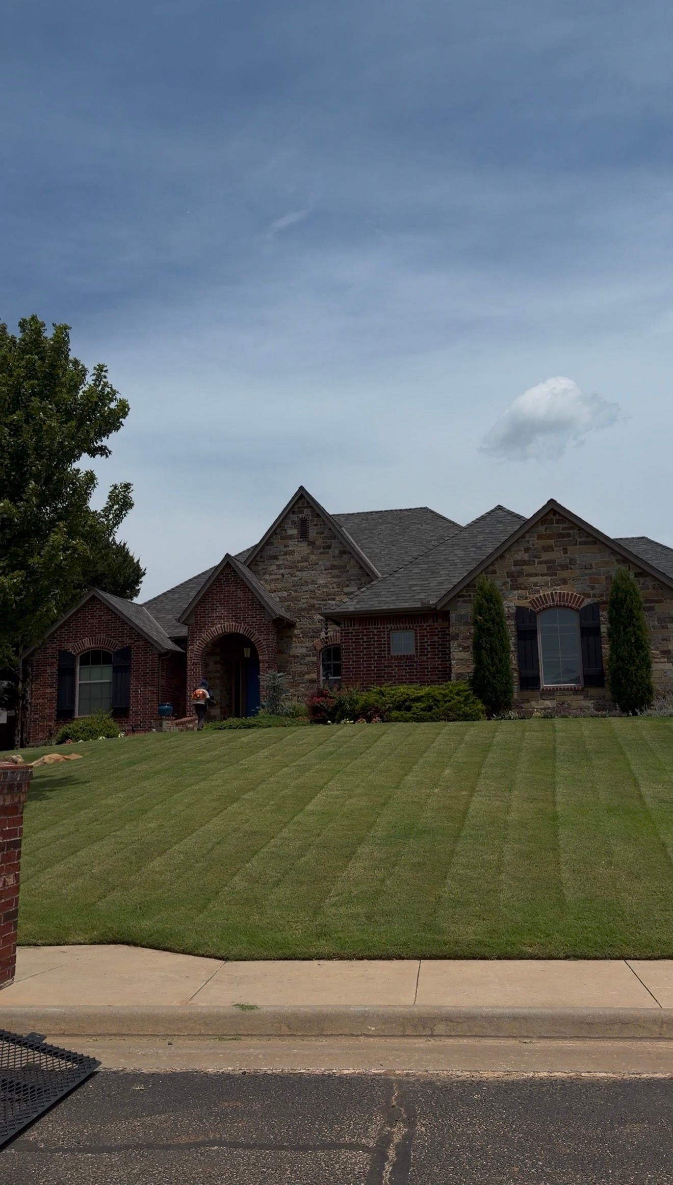 Brick suburban house with gray roof on a sloped lawn under a cloudy blue sky