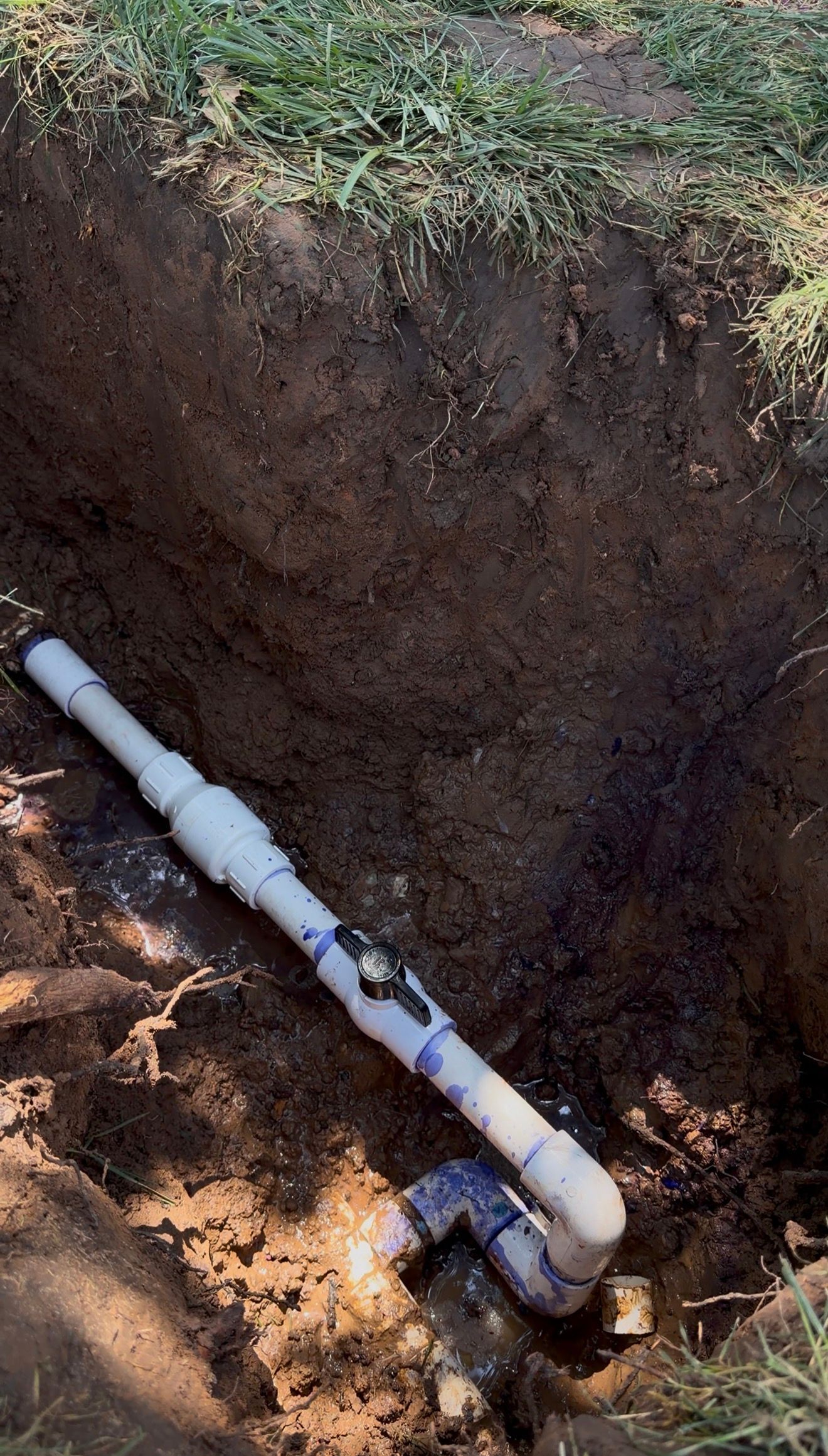 Broken white PVC pipe in a dirt trench beside exposed soil and grass.